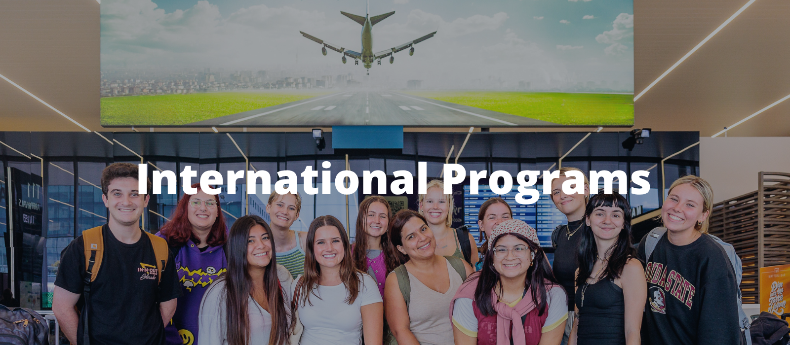A picture of students in an airport posing together with a picture above them of a plane taking off with the words "International Programs" overlaid on top of the photo.