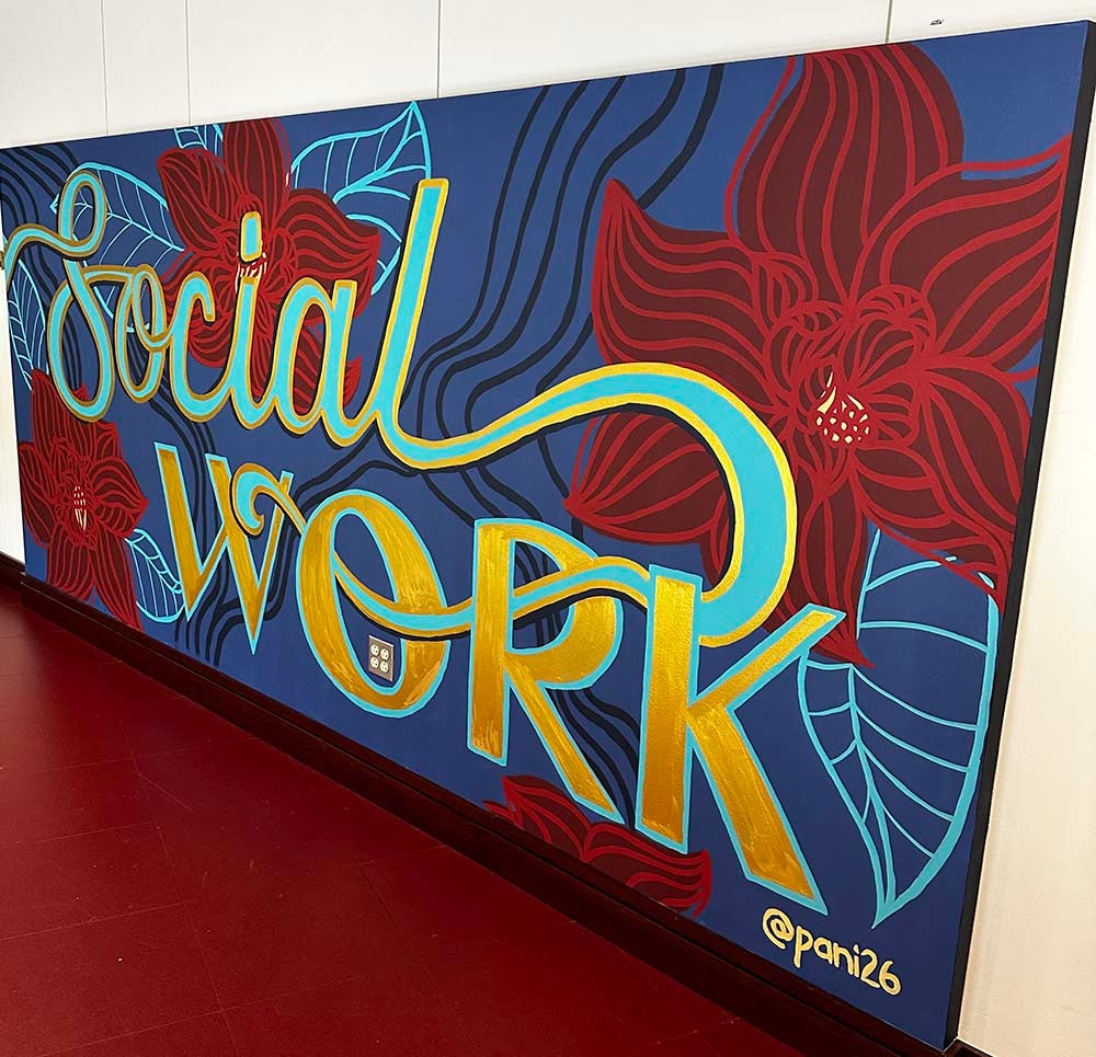 "Painted mural with the words “Social Work” in large gold and light blue lettering on a dark blue background. Red flowers, light blue leaves, and flowing abstract lines surround the text. The artwork is displayed on an interior wall above a red floor and includes the artist signature “@pani26” in the lower right corner."
