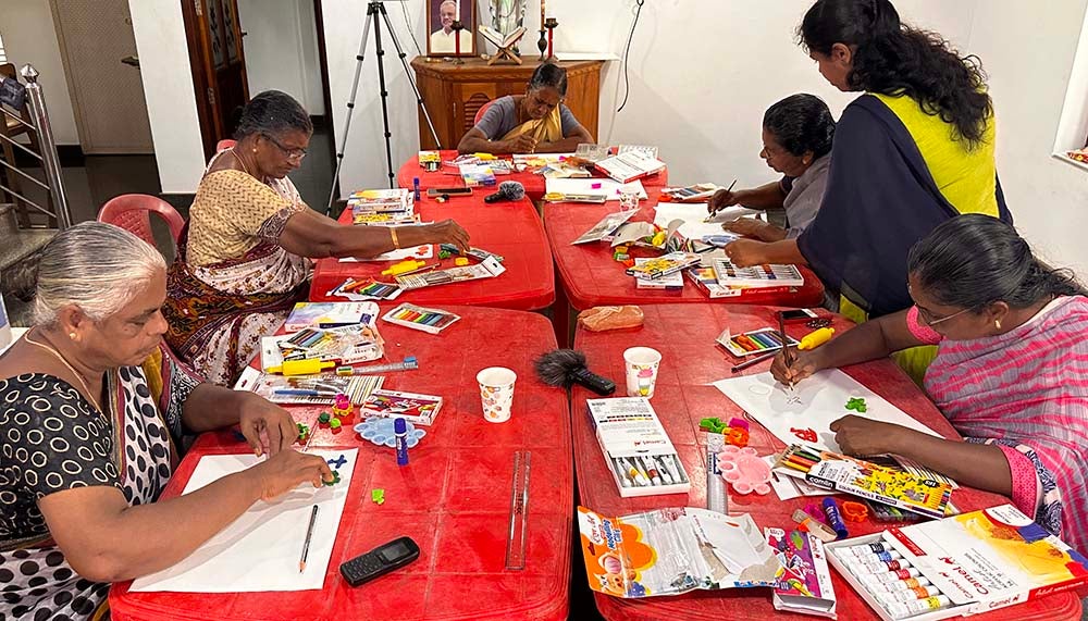 "Kerala residents participating in an art envisioning activity at bright red tables creating collages and art pieces surrounded by art supplies"