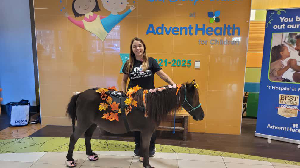 "Fabiana Rocha with a dark miniature horse dressed in fall leaves at AdventHealth hospital"