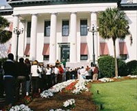 "An image of the Old Florida Capitol Building with columns and red and white striped awnings. People gather in front of the building and are bordered by white and red flowers and a green lawn."
