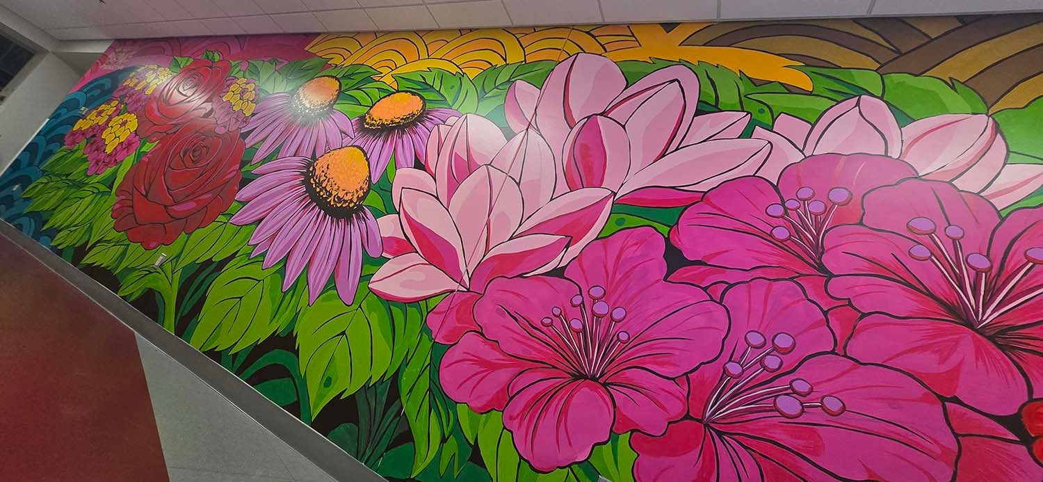 "Large floral mural painted along an interior hallway wall, featuring oversized pink blossoms, magnolia-like flowers, red roses, purple coneflowers, and green leaves. The colorful artwork spans the length of the corridor with pink, green, blue, and gold abstract background patterns. A ramp runs alongside the mural under overhead ceiling lights."