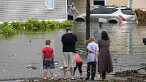 "Photo of two adults and three children with their back to the view looking at a flooded street, home and submerged car."