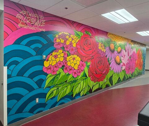 Colorful mural painted along an indoor hallway wall featuring large flowers in shades of red, pink, purple, yellow, and orange with green leaves. Blue wave-like patterns and a pink sky create the background. Artist signature text appears in the upper left corner. The hallway has overhead lighting and a red floor.  Provide your feedback on BizChat