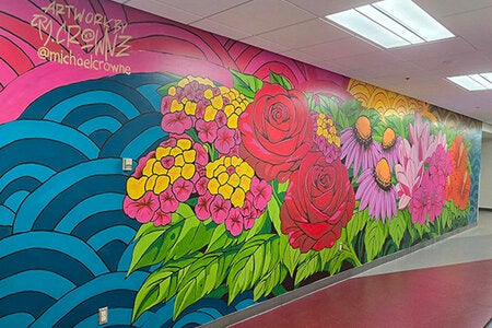 Colorful mural painted along an indoor hallway wall featuring large flowers in shades of red, pink, purple, yellow, and orange with green leaves. Blue wave-like patterns and a pink sky create the background. Artist signature text appears in the upper left corner. The hallway has overhead lighting and a red floor. 